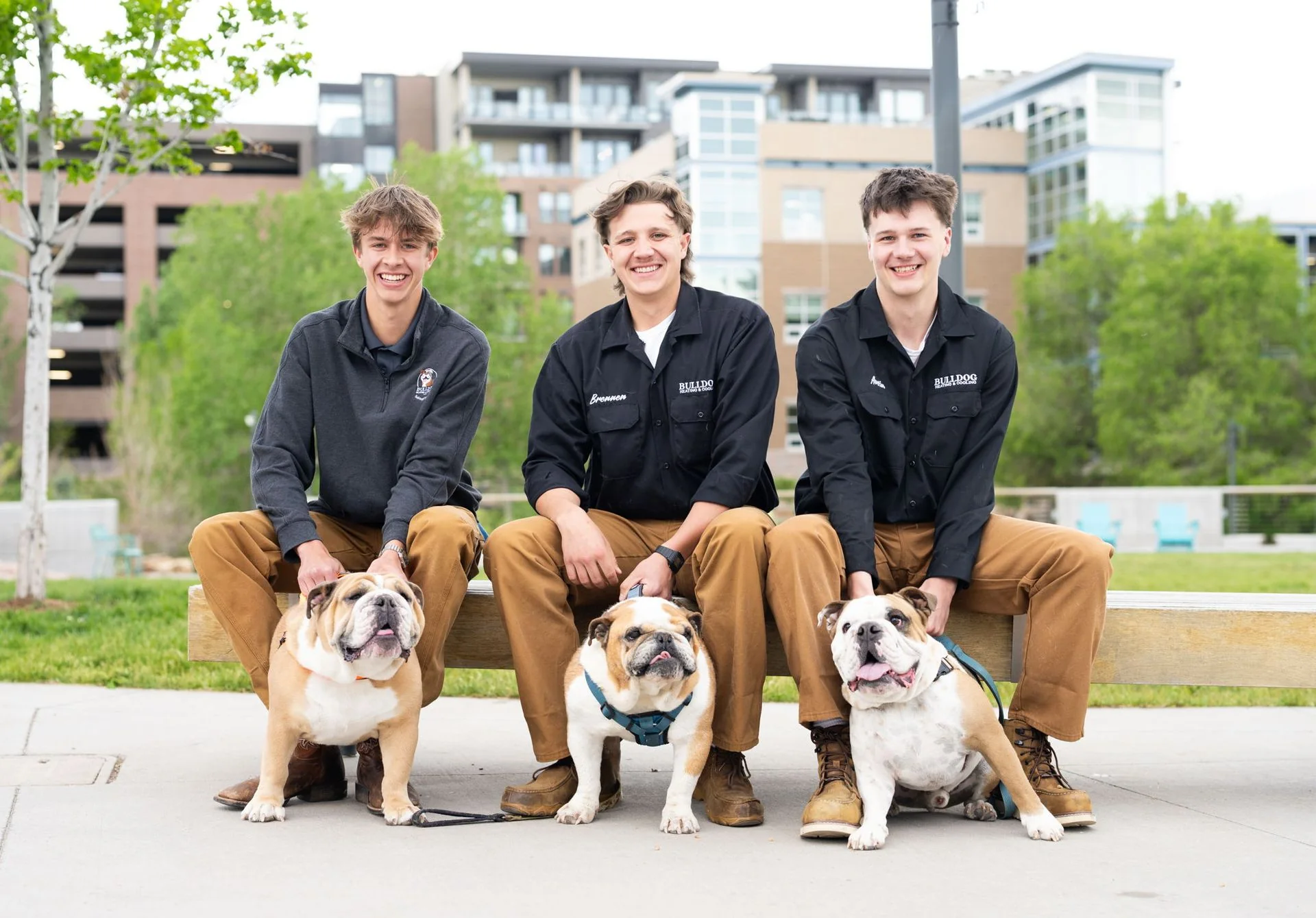 Three young men in black and tan uniforms sit on a bench with three bulldogs in an urban park setting